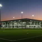 Um centro de treinamento de futebol moderno no Brasil ao pôr do sol, destacando a arquitetura avançada e campos iluminados, simbolizando o futuro e os investimentos em infraestrutura esportiva. Um jovem jogador observa a estrutura com esperança.