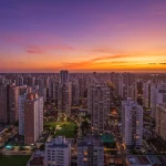 Vista aérea de uma cidade brasileira moderna ao pôr do sol, com muitos edifícios residenciais e áreas verdes, refletindo crescimento urbano.