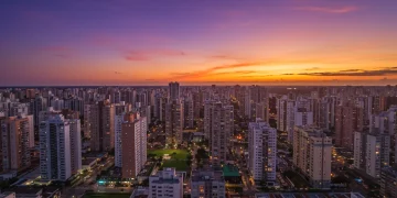 Vista aérea de uma cidade brasileira moderna ao pôr do sol, com muitos edifícios residenciais e áreas verdes, refletindo crescimento urbano.