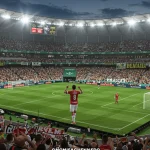 Estádio de futebol brasileiro lotado com jogadores e torcedores celebrando, destacando placas de publicidade e uniformes com logotipos de patrocinadores.