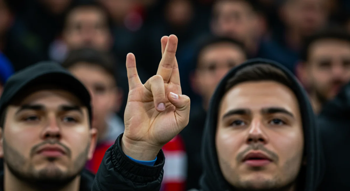 Mão de torcedor realizando um gesto supersticioso durante um jogo de futebol decisivo.