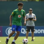 Jovem jogador de futebol brasileiro em campo, observando o futuro com o apoio de um agente.