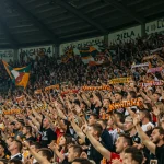 Torcedores vibrantes cantando em um estádio lotado, bandeiras e fumaça colorida ao fundo. A cena reflete a paixão e a energia das torcidas organizadas no futebol brasileiro.